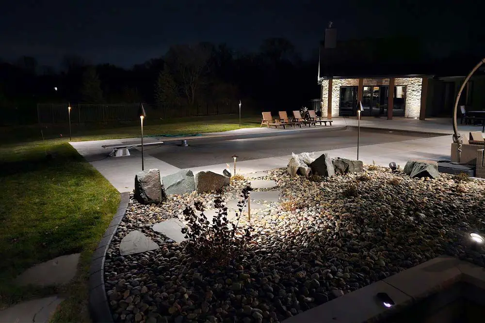 Nighttime backyard patio with landscape lighting highlighting a rock garden, stepping stones, and modern seating area.