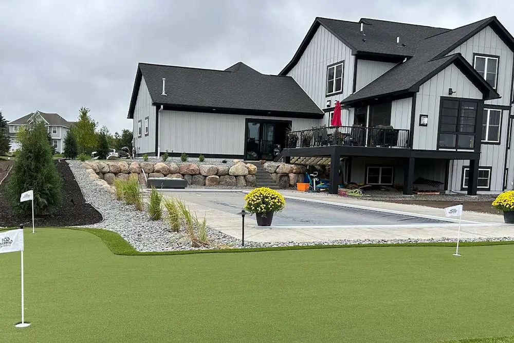 Modern backyard with artificial turf putting green, concrete patio, stone boulder retaining wall, and gray farmhouse-style home with raised deck and potted yellow flowers.