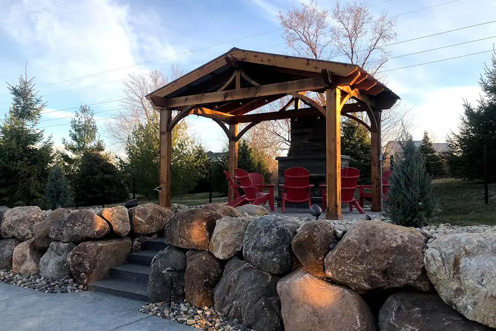 Backyard outdoor living space with a wooden pavilion, red Adirondack chairs, stone fireplace, and large boulder retaining wall surrounded by evergreen trees.